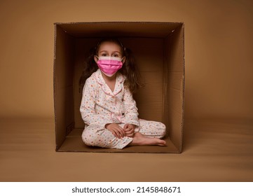 Cute little Caucasian girl with two ponytails in pajamas wearing a pink medical protective mask sits inside a cardboard box, looks at camera, isolated over beige background with copy ad space