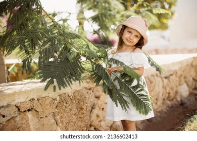 Cute Little Caucasian Girl In A White Dress And Straw Hat Stands Near A Green Tropical Tree In A Luxury Resort. The Concept Of Summer Vacation.