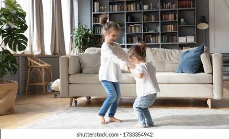 Cute Little Caucasian Girls Sisters Hold Hands Dancing In Cozy Living Room Together, Small Preschooler Children Play Have Fun At Home, Jumping And Swirling Engaged In Funny Childish Activity