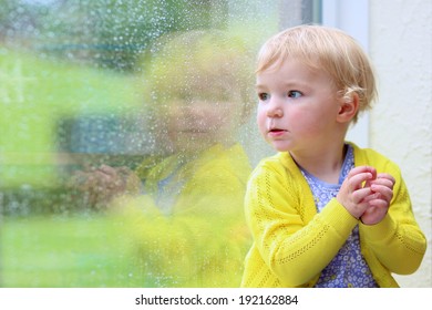 Cute Little Child, Blonde Curly Toddler Girl In Colorful Casual Outfit Sitting Indoors On A Rainy Day Looking Through Window With Garden View