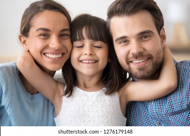 Cute little child daughter embracing caring parents mom and dad looking at camera, preschool kid girl embracing loving mother and father posing together, smiling family of three headshot portrait