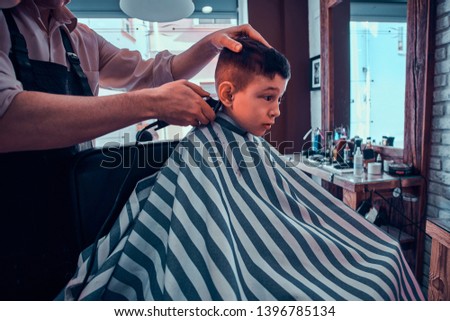 Cute little child is getting trendy haircut from barber at busy barbershop.