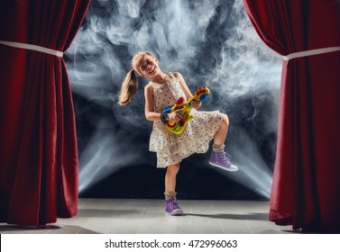 Cute Little Child Girl Playing Guitar On Stage. Kid Dreams Of Becoming A Rock Musician.