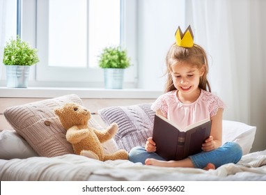 Cute Little Child Girl Reading A Book In The Bedroom. Kid With Crown Sitting On The Bed Near Window.