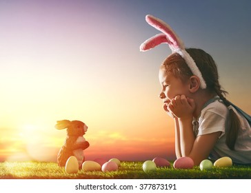 Cute little child girl wearing bunny ears on Easter day. Girl hunts for Easter eggs on the lawn. Girl with Easter eggs and bunny in the rays of the setting sun. - Powered by Shutterstock