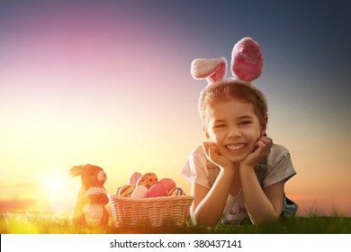 Cute little child girl wearing bunny ears on Easter day. Girl hunts for Easter eggs on the lawn. Girl with Easter eggs and bunny in the rays of the setting sun. - Powered by Shutterstock