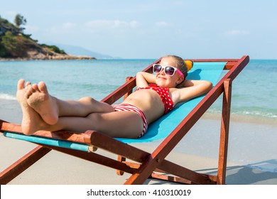 Cute little child in red swimsuit and white sunglasses smile and enjoying on a sun lounger rest on the beach chair on tropical sandy beach sea shore. Sunbathing and leisure on sunny day.