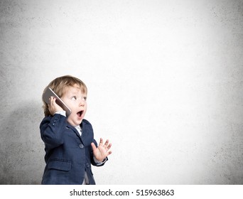 Cute Little Child Is Talking On His Smartphone While Standing In His Business Suit Near A Concrete Wall. Mock Up