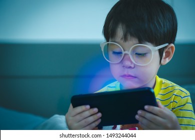 Cute Little Child Watching Cartoon On Smartphone In The Dark Room. Dangers Of Blue Light Damage His Eyes. Handsome Little Boy Can Get Age Related Macular Degeneration Of Blue Light, He Wear Eyeglasses