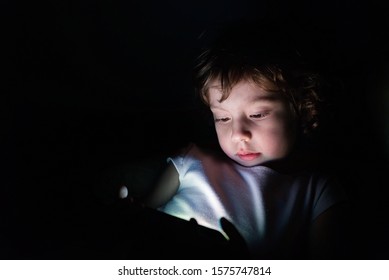 Cute Little Child Watching Cartoon On Smartphone In The Dark Room. Dangers Of Blue Light Damage His Eyes.  Little Girl Can Get Age Related Macular Degeneration Of Blue Light.
