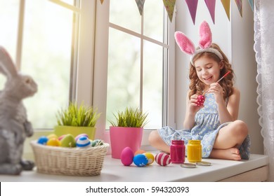 Cute Little Child Wearing Bunny Ears. Girl Sitting On The Window. Kid Is Painting Eggs.