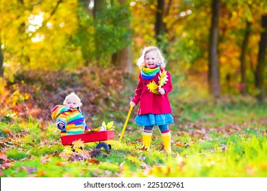 Cute Little Children, Adorable Toddler Girl And A Funny Baby Boy, Brother And Sister, Playing In A Sunny Autumn Park With A Wheel Barrow And Colorful Leaves