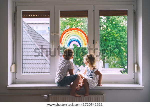 Cute little children on background of painting\
rainbow on window. Photo of kids leisure at home, safety joy\
symbol, happy childhood. Positive visual support during quarantine.\
Family Art Background