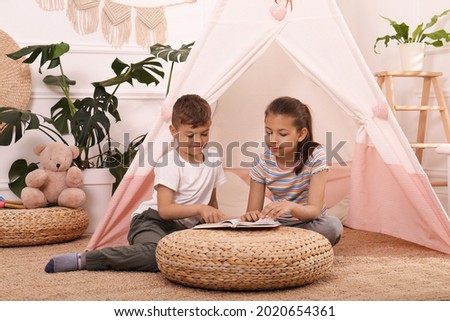 Cute little children reading book near toy wigwam at home