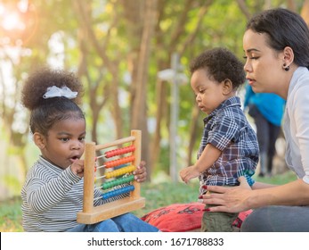 Cute Little Dark Skinned, Mix Race African Toddler Girl Playing With Wooden Educational Toy, Sitting With Asian Mom And Baby Brother In The Park At Picnic. Family Quality Bonding Time Together.