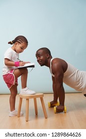 Cute Little Daughter Fitness Training Her Handsome Black Young Father. She Is Making Notes In Her Paper Notebook. Child Role Play. He Looks At Her For Approval.