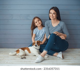 Cute little daughter and her beautiful young mother are looking at camera and smiling while posing with their dog