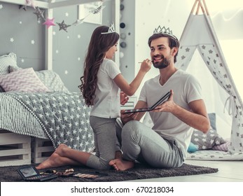 Cute Little Daughter And Her Handsome Young Dad In Crowns Are Playing Together In Child's Room. Girl Is Doing Her Dad A Makeup