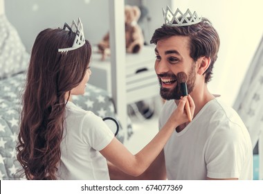 Cute Little Daughter And Her Handsome Young Dad In Crowns Are Playing Together In Child's Room. Girl Is Doing Her Dad A Makeup