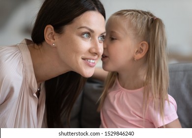 Cute Little Daughter Telling Secret To Smiling Young Mother, Whispering In Ear Close Up, Sitting On Couch At Home, Adorable Preschool Child Sharing Rumor With Happy Mum, Gossiping, Having Fun