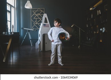 Cute little dreamer boy posing with a helmet at home, pretending to be an astrounaut, cardboard space rocket in the background