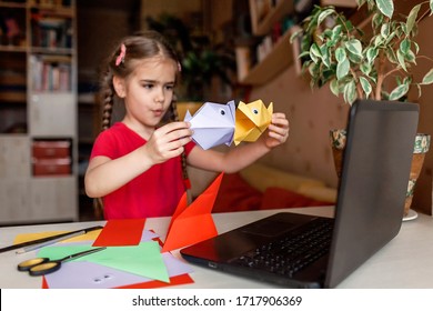 Cute little elementary schoolgirl doing origami fish with folded color paper looking video on laptop, online workshop, kids at-home activity, creativity and distant education, focus on fish