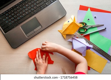 Cute little elementary schoolgirl doing origami fish with folded color paper looking video on laptop, online workshop, kids at-home activity, creativity and distant education