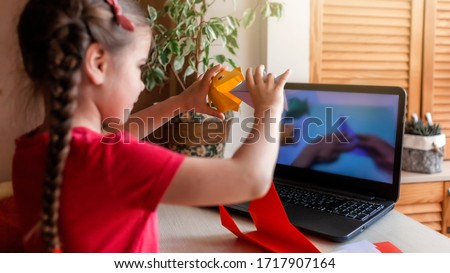 Cute little elementary schoolgirl doing origami fish with folded color paper looking video on laptop, online workshop, kids at-home activity, creativity and distant education, focus on hands