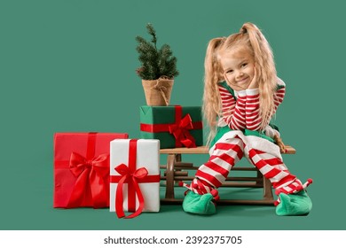 Cute little elf with Christmas gifts and sled on green background