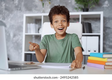 Cute little esxcited schoolboy sitting at the table in the classroom and pointing with index finger at the laptop computer monitor andlooking to the camera with surprised expression                    - Powered by Shutterstock