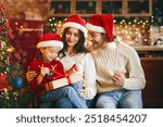 Cute little festive girl openng xmas gifts on Christmas eve with mom and dad, decorated kitchen interior, copy space