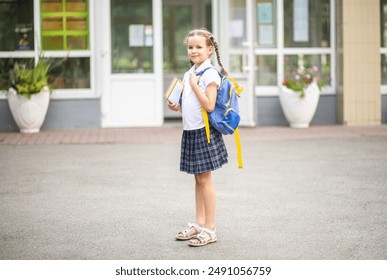 A cute little first-grader girl stands near the school with a blue school backpack. The child goes to first grade. Back to school. A girl in the school yard before her first lesson.  - Powered by Shutterstock