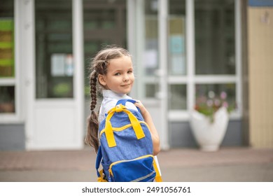 A cute little first-grader girl stands near the school with a blue school backpack. The child goes to first grade. Back to school. A girl in the school yard before her first lesson.  - Powered by Shutterstock