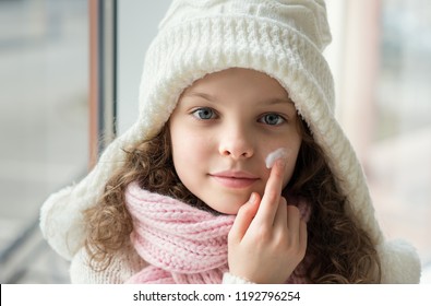 Cute Little Girl Applying Protecting Cream To Her Face. Winter Skin Protection Concept.   Portrait Of Smiling Kid In Winter Clothes.