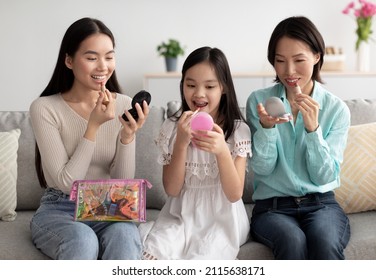 Cute Little Girl Asian With Her Mom And Granny Applying Makeup On Couch At Home. Happy Multi Generation Family Using Cosmetics, Having Domestic Spa Day In Living Room