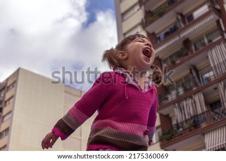 Cute little girl with bangs and pigtails happily screaming. She wears a pink striped sweater and a flowered shirt. Buildings and a cloudy sky in the background