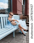 Cute little girl in beautiful blue poofy dress and sunglasses drinking cocktail sitting on a bench on city street. Fashion kids