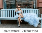 Cute little girl in beautiful blue poofy dress and sunglasses drinking cocktail sitting on a bench on city street. Fashion kids