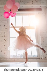 Cute little girl in beautiful dress is dancing at light sunny room with pink air balloons in hand.