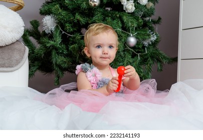 A cute little girl with a beautiful, perky smile, joyfully welcoming Christmas and the New Year by the ornate Christmas tree and its toys, in a dress and a beautiful interior. - Shutterstock ID 2232141193