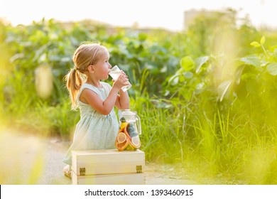 Cute Little Girl With Blonde Hair Drinking Lemonade Outdoor. Detox Fruit Infused Flavored Water, Cocktail In A Beverage Dispenser With Fresh Fruits