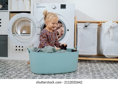 Cute Little Girl With Blonde Hair, Small Child Sits In Bathroom, Laundry Room By Large Bowl Full Of Clothes, Daughter Throws Things Into Washing Machine, Helps Mom With Daily Chores