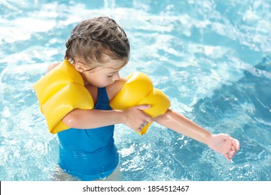 Cute Little Girl In Blue Swimwear Putting On Yellow Inflatable Safety Sleeves On Arms While Going To Have Swimming Lesson In Transparent Water