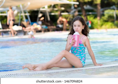 Cute little girl in bright swimsuit sitting with a cocktail by the pool on sunny summer day. Family vacation concept
