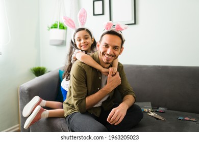 Cute Little Girl With Bunny Ears Hugging Her Happy Dad From Behind And Smiling While Making Eye Contact. Latin Daughter Putting Makeup And Playing With Her Father