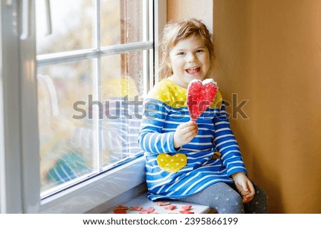Cute little girl by window holding selfmade heart as gift for Valentine's day, Mother's day or birthday. Adorable happy smiling child indoors.