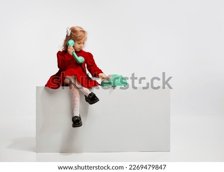 Cute little girl, child in beautiful red dress playing with retro phone against grey studio background. Concept of childhood, game, friendship, activity, leisure time, retro style, fashion.