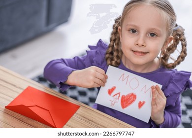cute little girl child is holding in hands handmade greeting card and red heart shape   sight Mom that she prepared for her mother as present for mother's day womans day 