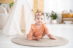 A Cute Little Girl Child In A Pink Bodysuit Crawls On A Rug In The Living Room Of The House Against The Background Of A Wigwam, Looks At The Camera, Smiles