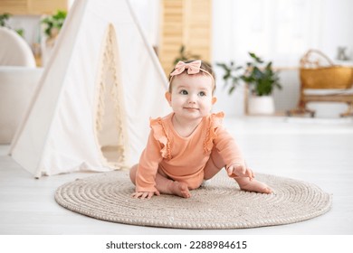 a cute little girl child in a pink bodysuit crawls on a rug in the living room of the house against the background of a wigwam, looks at the camera, smiles - Shutterstock ID 2288984615